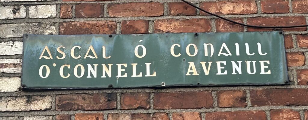 Photograph of street name plate for O’Connell Avenue, with the name in Irish and in English, white text on a green background.