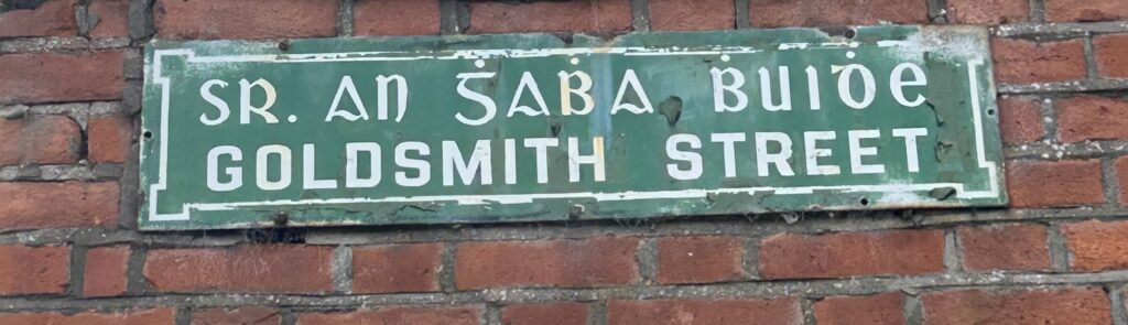 Photograph of street name plate for Goldsmith street, in Irish and in English, in white text on a green background.