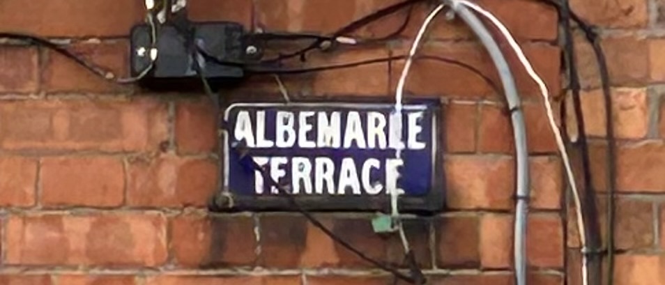 Photograph of street name plate for Albemarle Terrace. The text is white on a blue background, and the sign is affixed to the red brick wall of a house.