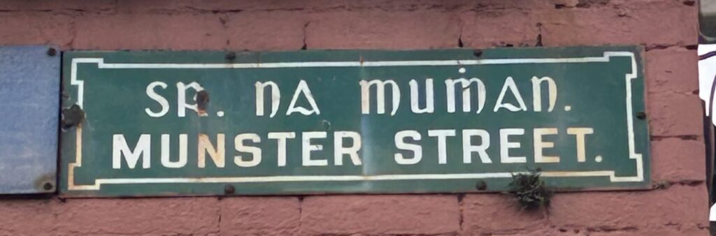 Photograph of street name plate for Munster Street, in Irish and in English, in white text on a green background.
